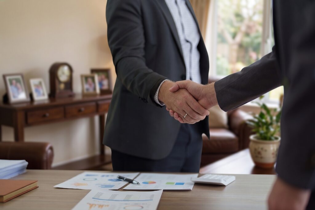 Two men in suits shaking hands across a wooden desk with financial charts, a calculator, and paperwork in a home setting.